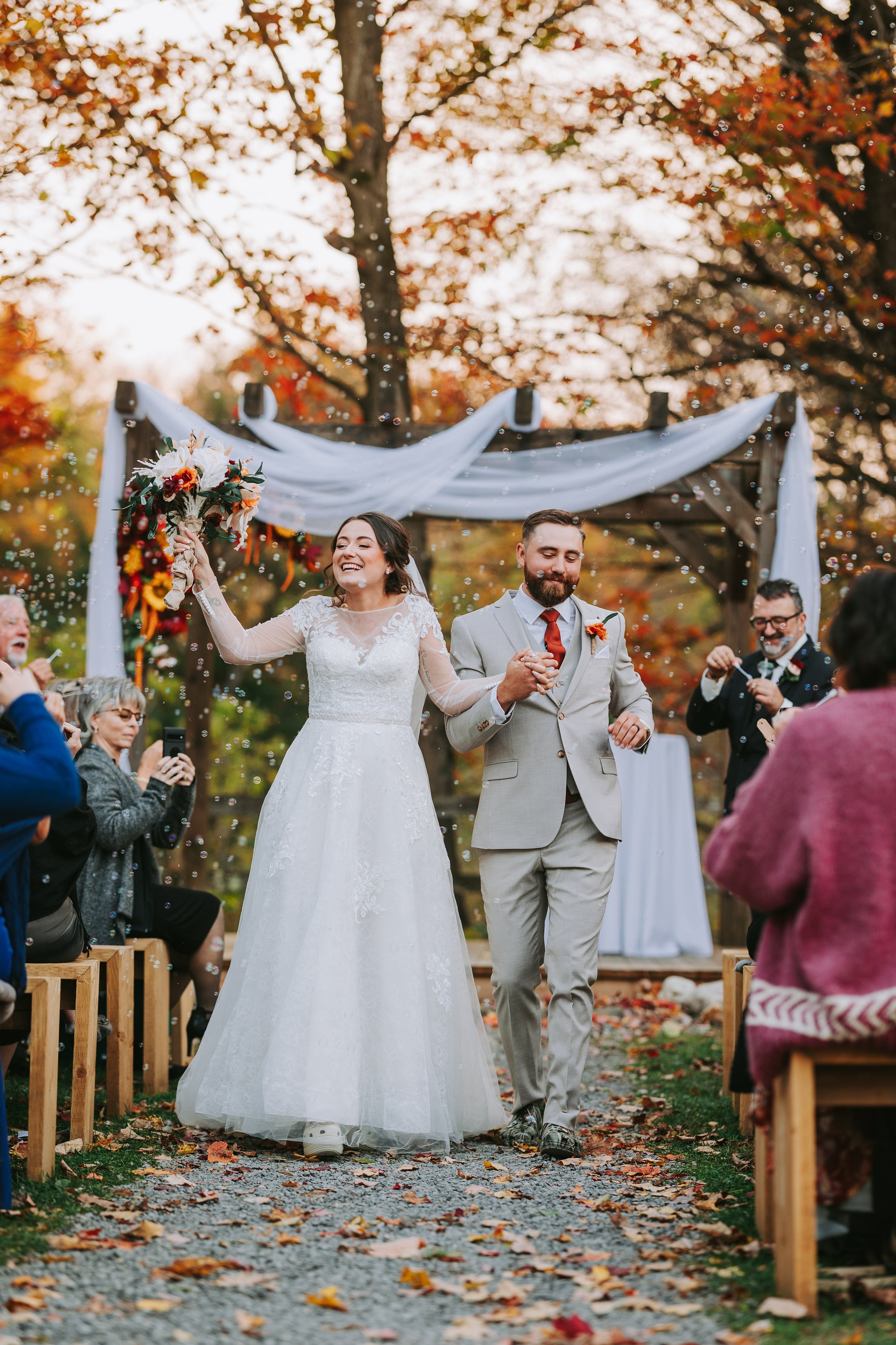 A newlywed couple walks down an outdoor aisle holding hands, celebrating their wedding, with guests taking photos and throwing confetti amid fall foliage.