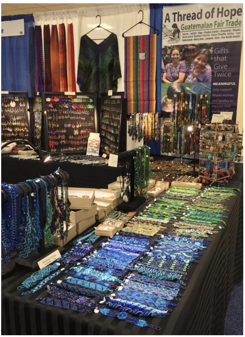 A woman smiling and sitting on the ground outdoors, working with a loom to weave fabric.