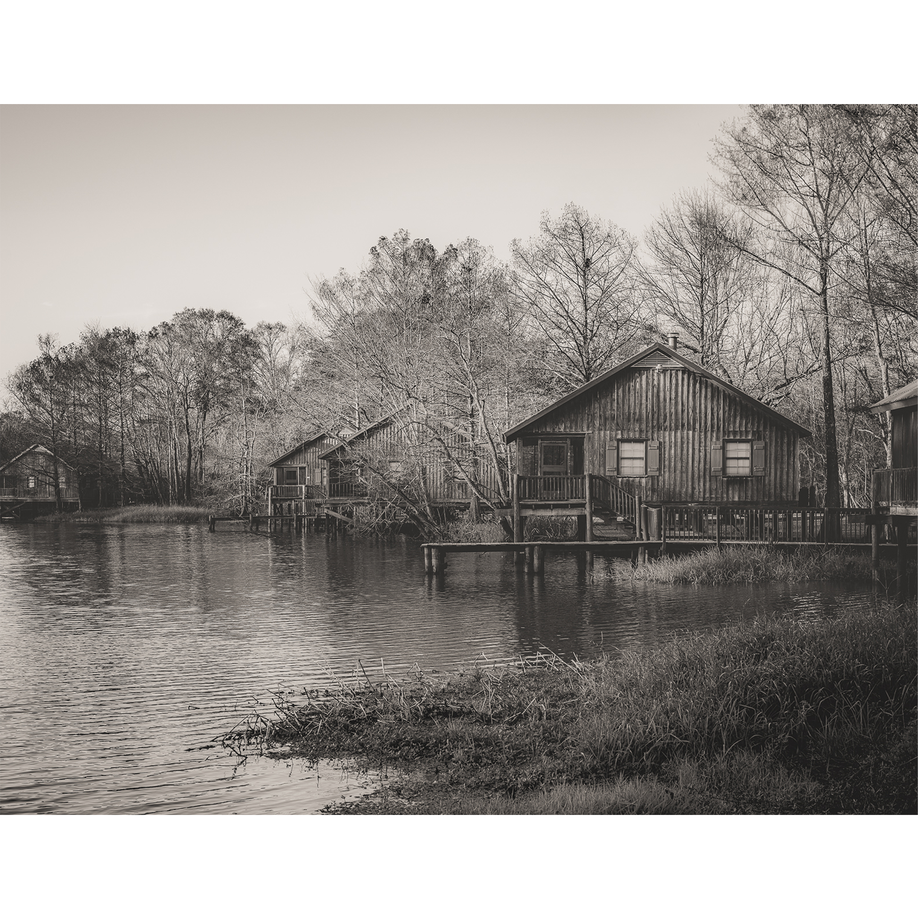 Monochrome photograph of rustic wooden cabins lining the calm waters of a Louisiana bayou, framed by leafless winter trees.