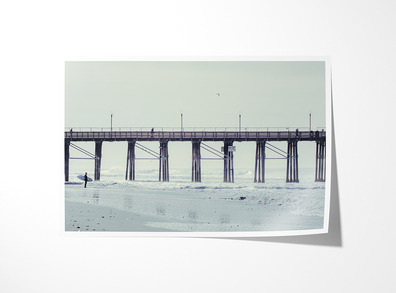 Framed fine art print of a surfer pausing before entering the ocean, dwarfed by the quiet symmetry of a pier stretching out over the tide.