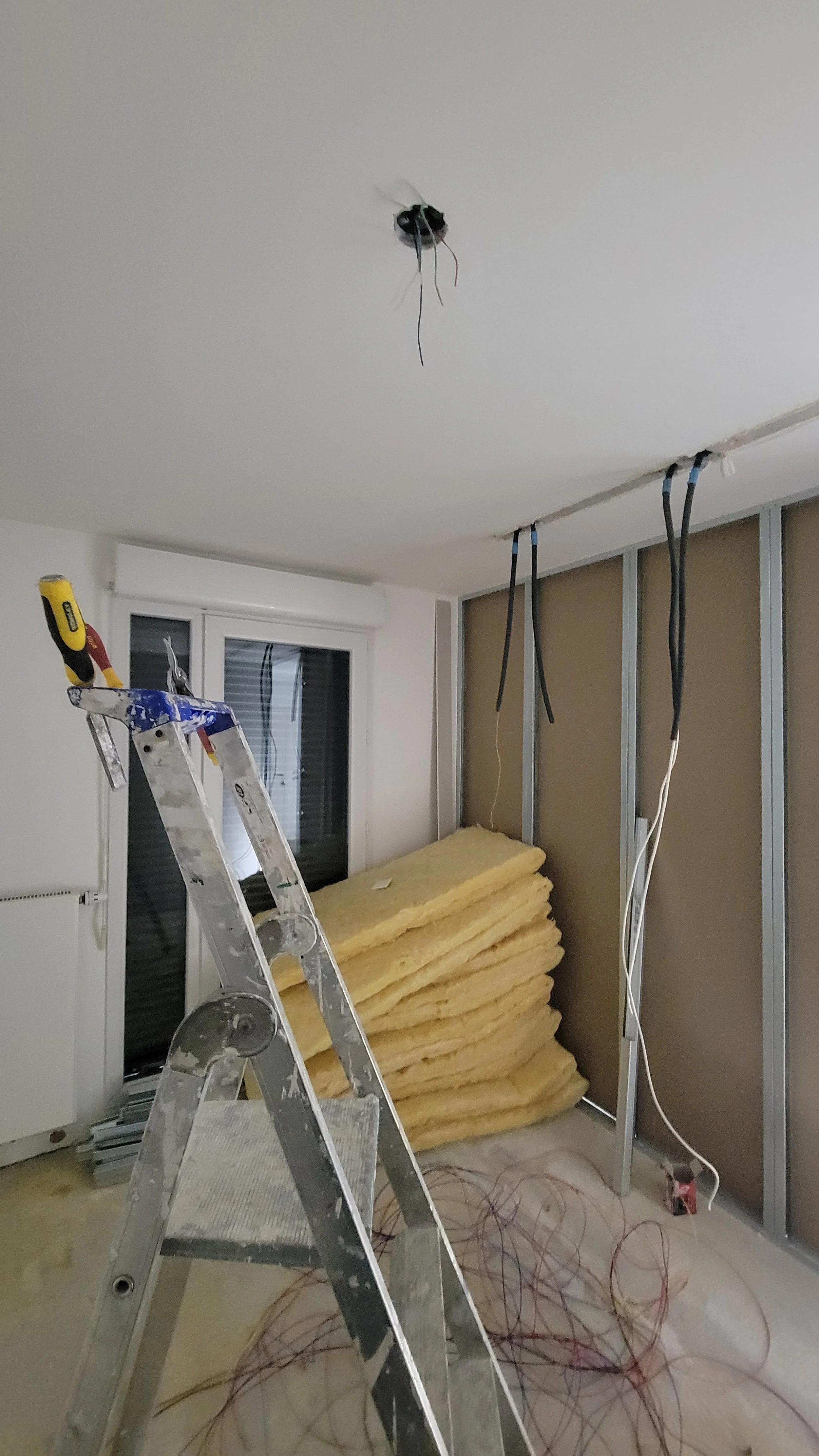 Une pièce en rénovation avec un plafond en cours d’installation, des cosses électriques suspendues, une échelle, des matériaux isolants empilés et des câbles électriques au sol.