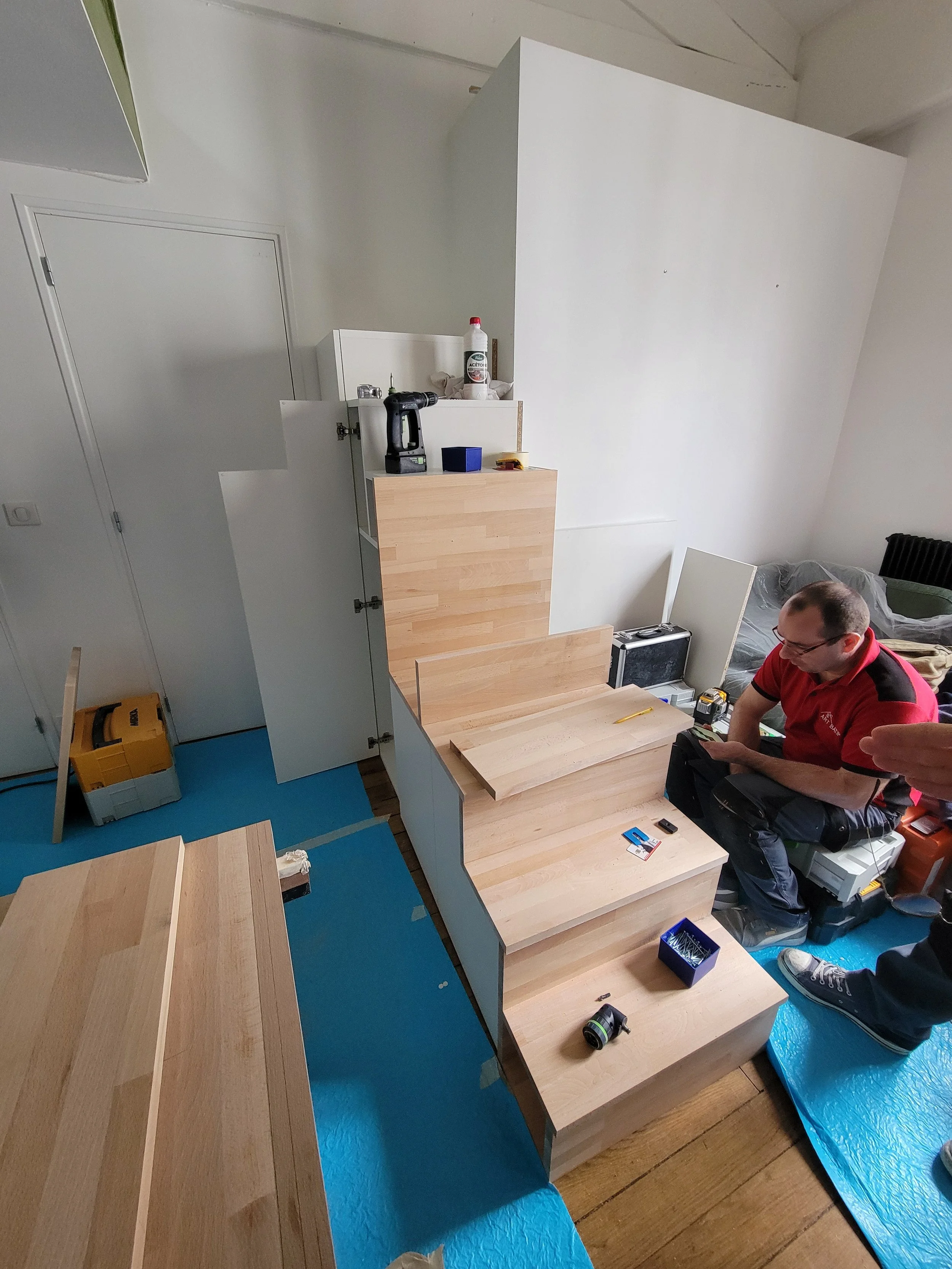 Une personne en train d'installer un escalier en bois dans une pièce. Le travailleur utilise des outils et des matériaux de construction, avec un sol recouvert d'une nappe bleue.
