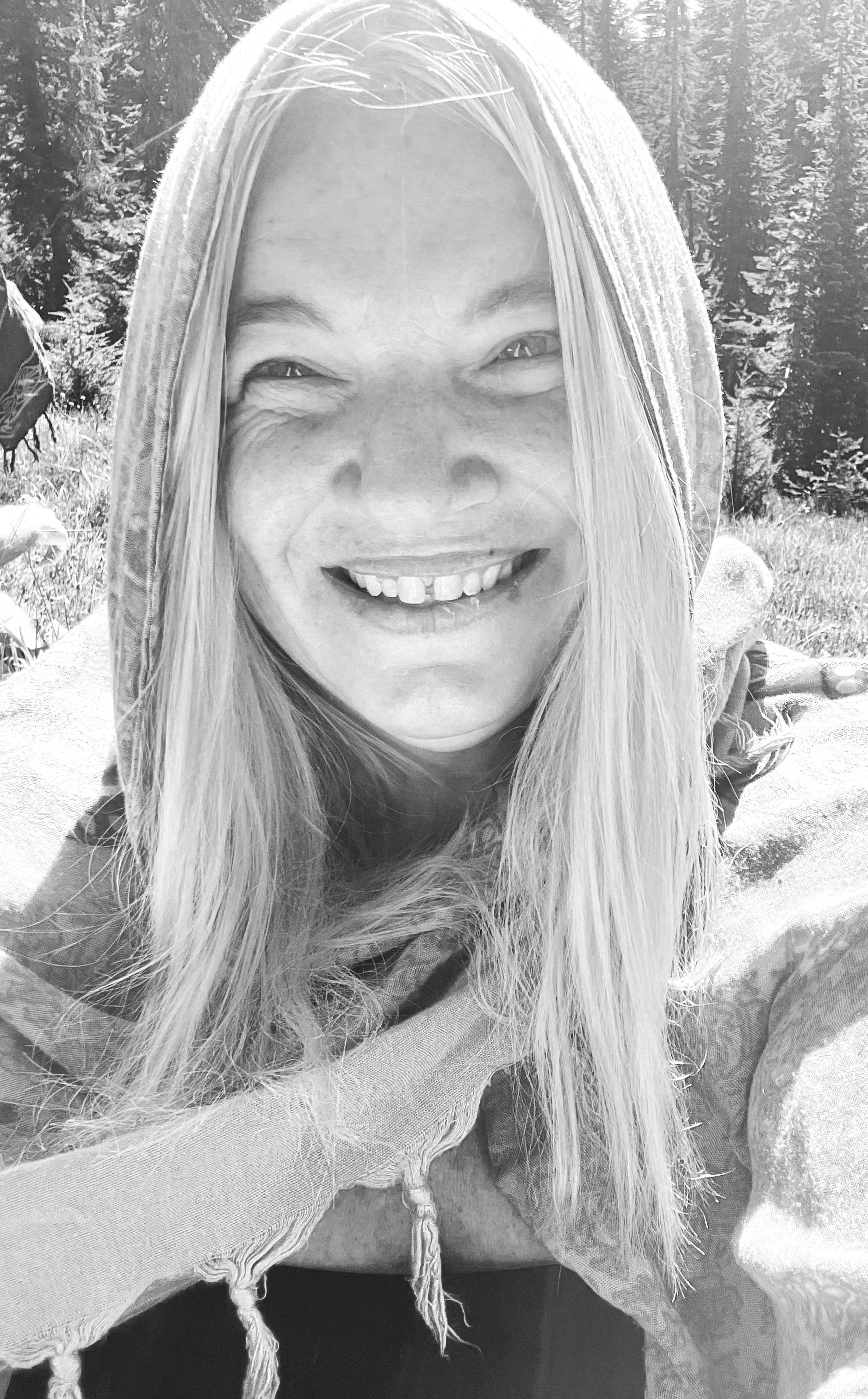 Black and white photo of a woman smiling outdoors with trees in the background, wearing a hooded jacket.