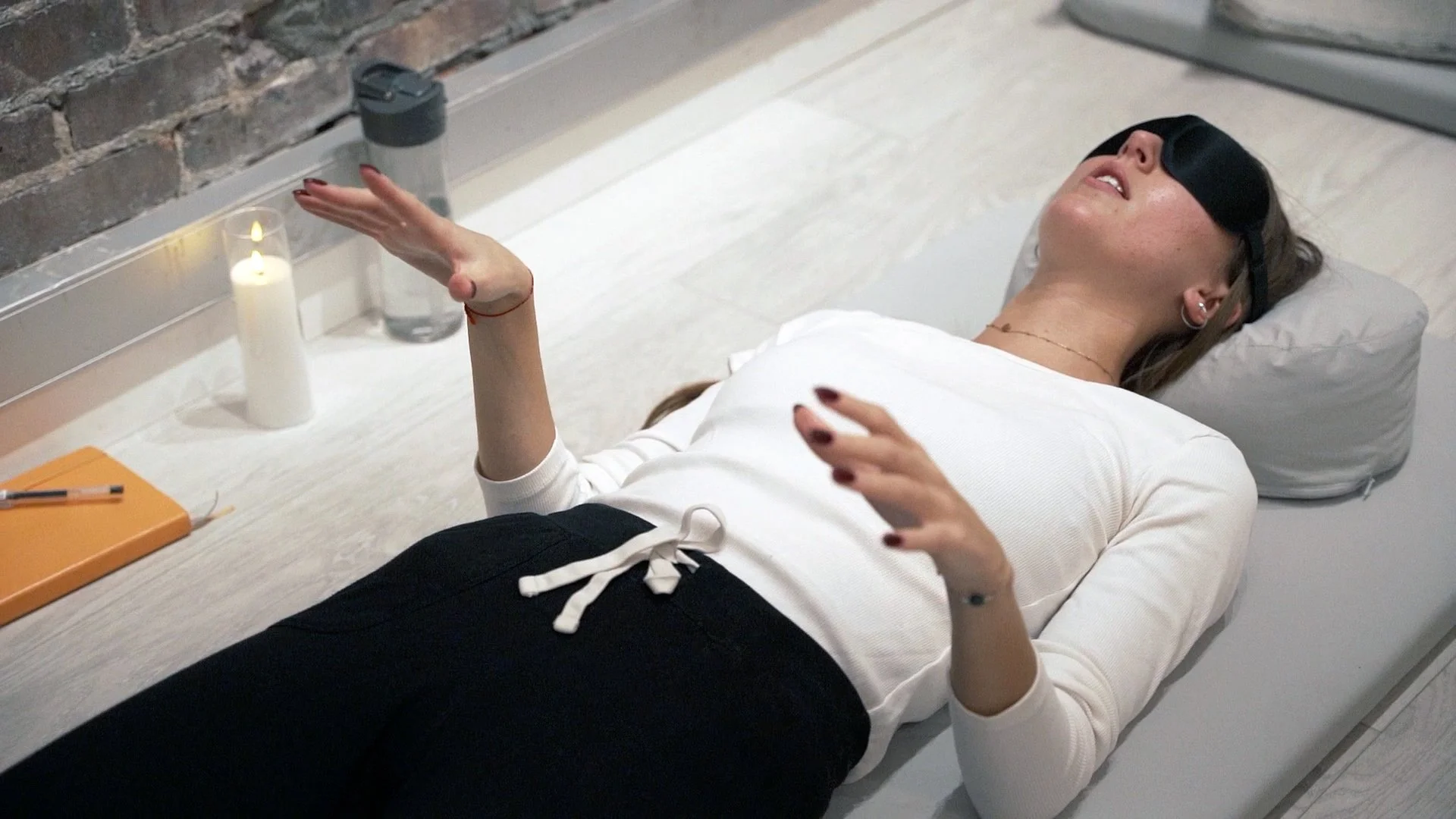 A woman lying on a bed with an eye mask, appearing relaxed, with a candle, water bottle, orange notebook, and pen on a nearby desk.