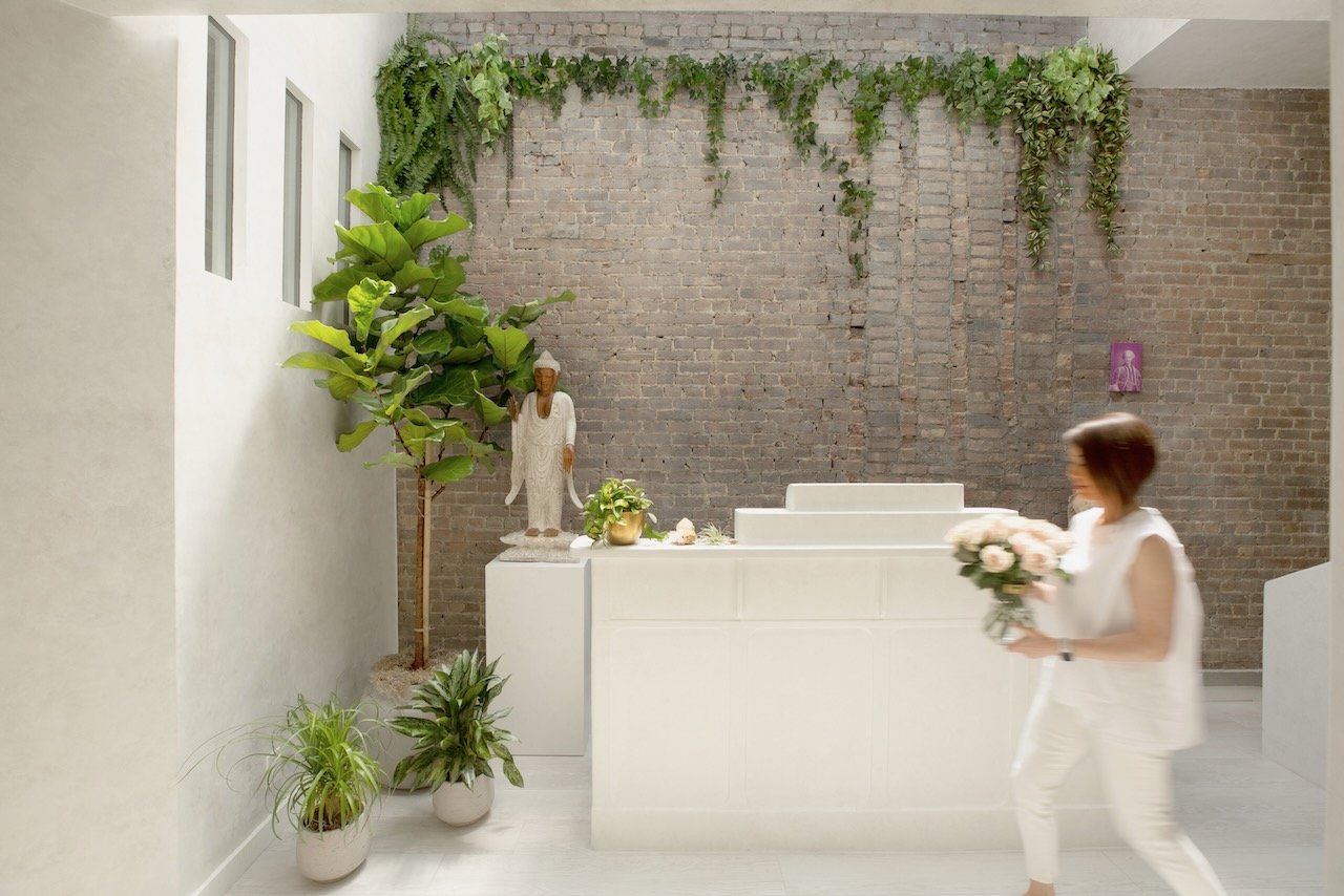 Woman holding a bouquet of flowers walking in a room with a white altar, potted plants, a brick wall, and a statue.