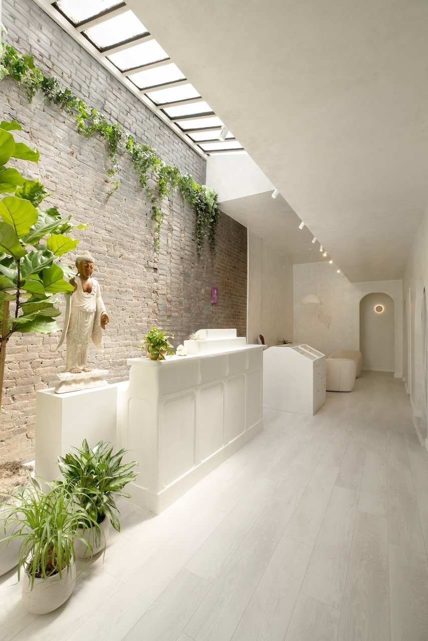 Minimalist interior with white and beige decor, potted plants, a statue, brick wall, skylight, and modern lighting.