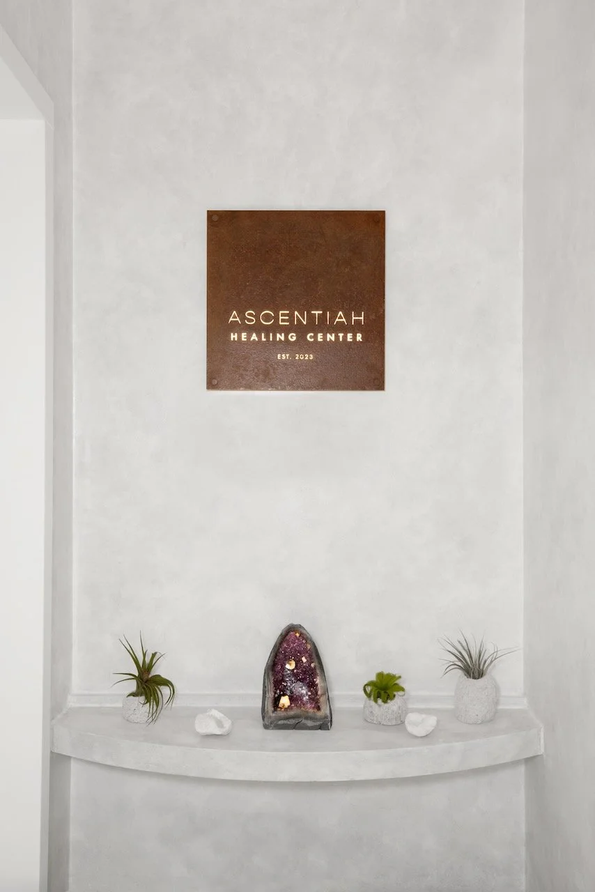 Decorative display shelf with three small potted plants and a geode, near a sign for Ascentiah Healing Center on a white wall.