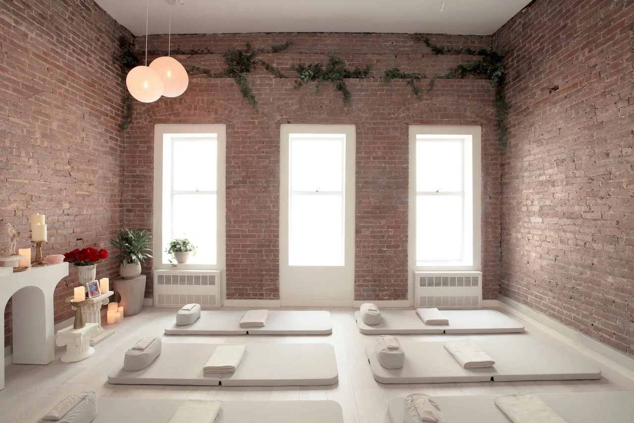A yoga or meditation studio with white mats, folded towels, and small pillows on a light wooden floor. The room has three tall windows with white frames, exposed brick walls, and minimal decor including potted plants, candles, and hanging spherical l