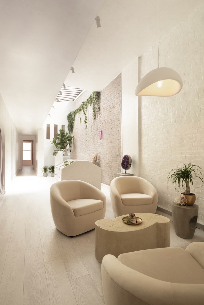 Bright, modern interior with cream-colored armchairs, a small oval table, and various potted plants against a white brick wall with greenery, under a hanging light fixture.