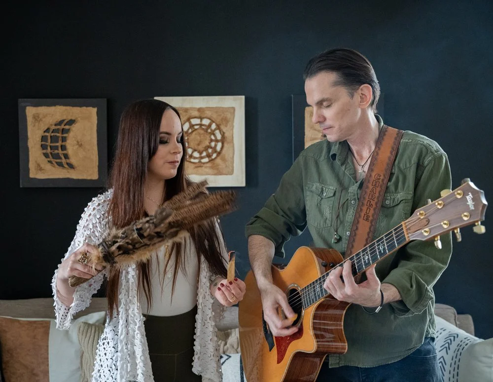 A man playing an acoustic guitar with a woman holding a fan and a feather accessory, standing in a room with framed artwork on a dark wall.