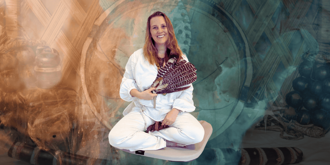 A woman with long red hair, smiling and sitting cross-legged, with hands in a prayer position, surrounded by percussion instruments including a large drum, a rattle, and a maraca, in front of a window showing an autumn landscape.