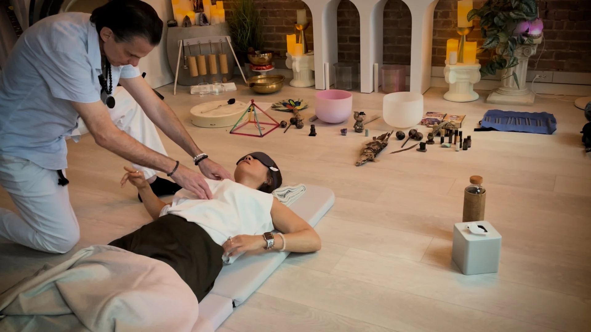 A person lying on a mat receiving CPR from another person in a room with spiritual and healing objects.
