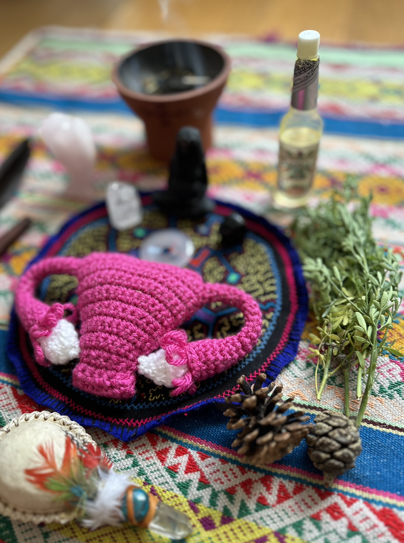 A colorful, patterned tablecloth with various spiritual and natural items, including a pink crocheted knot, pine cones, herbs, a small bottle, feathers, and stones.