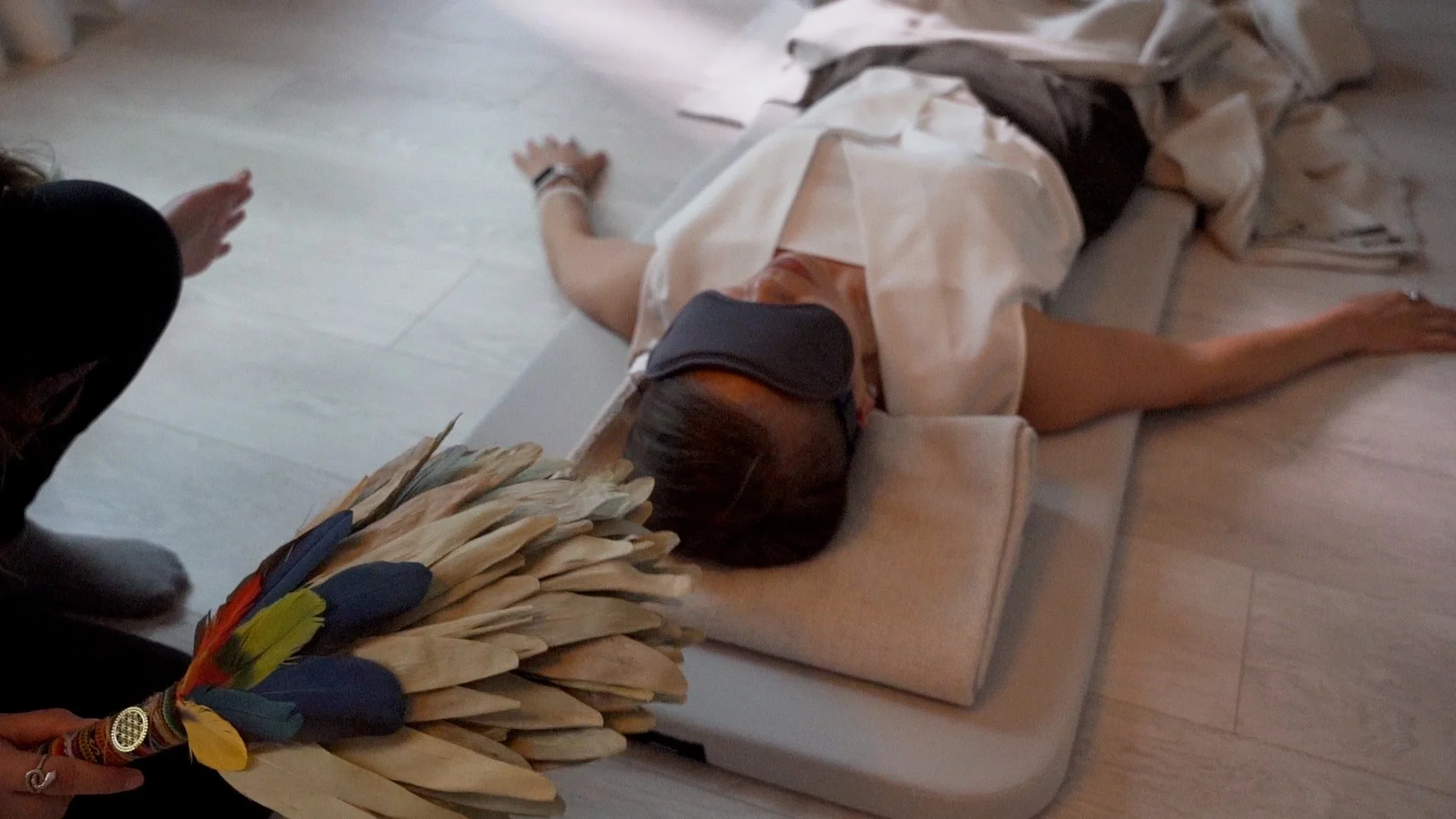 A person lying face down on a cushioned surface, wearing a face mask and traditional attire, with a large bundle of colorful feathers and sticks held by another person nearby.