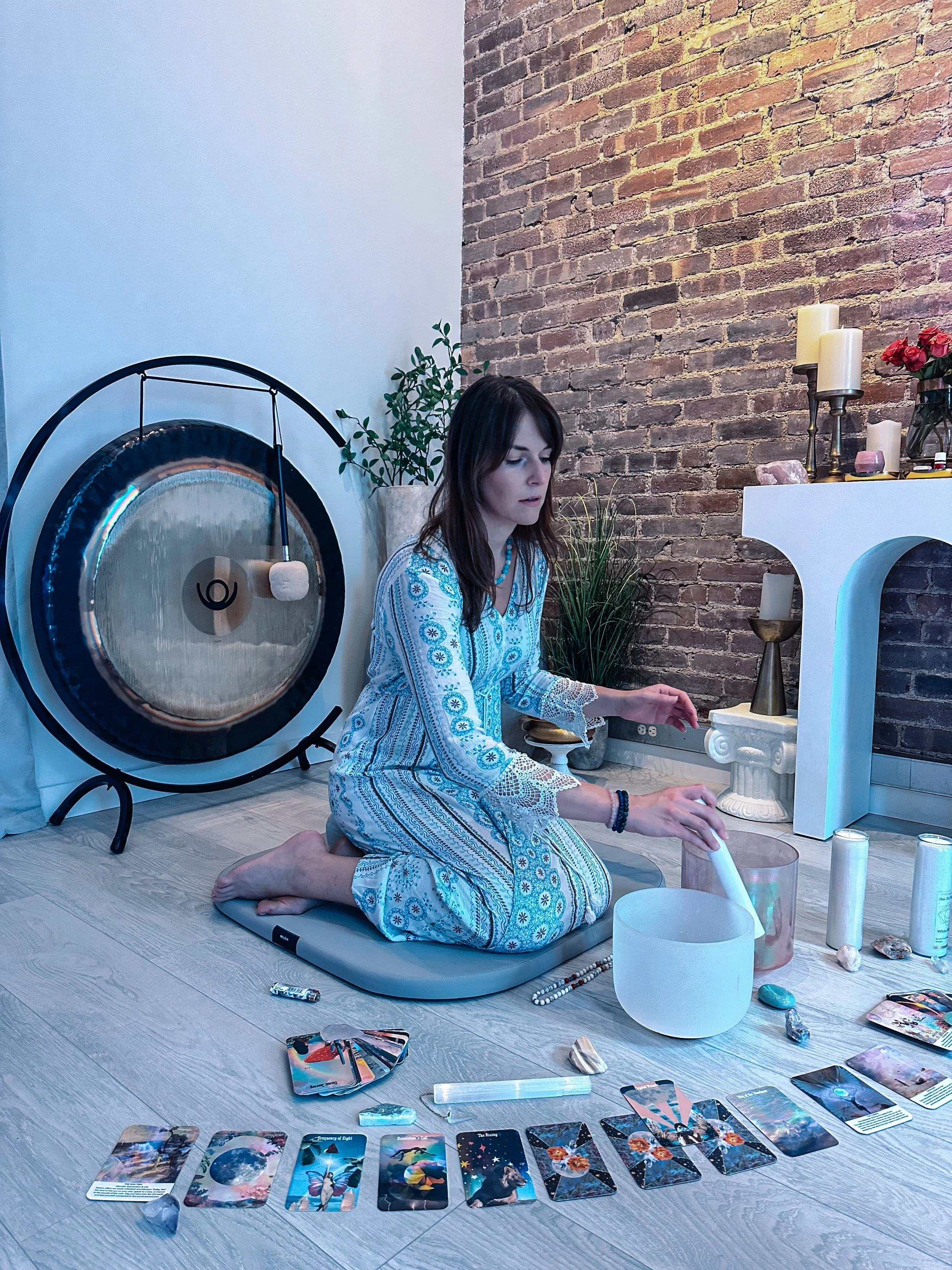 Woman in bohemian attire kneeling on floor surrounded by tarot cards, crystals, and candles, using a singing bowl, in a room with a brick wall, plants, and a large gong.