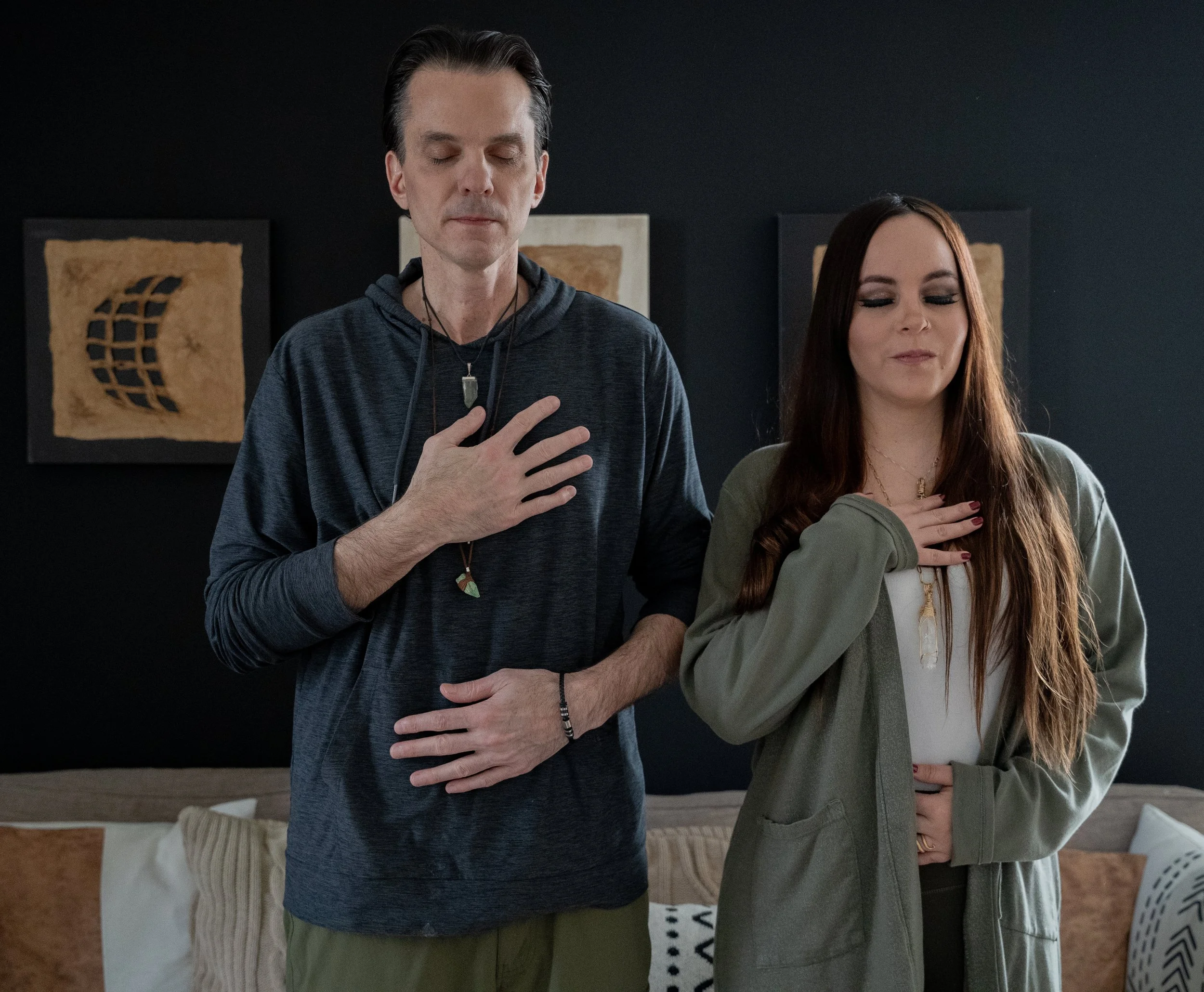 A man and woman standing side by side with their eyes closed, each with one hand over their chest, appearing to be in a moment of reflection or meditation in a cozy, modern living room.