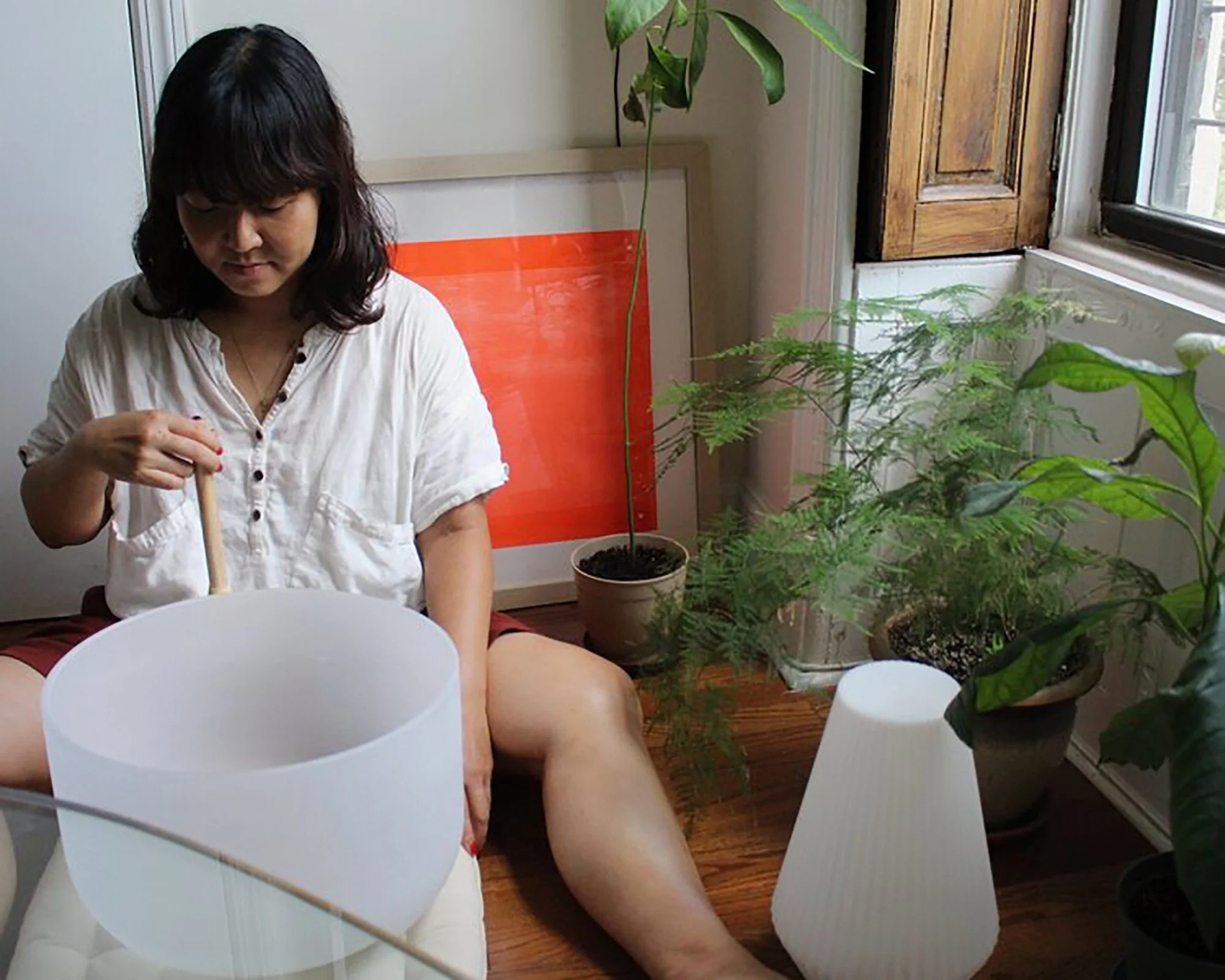 A woman sitting on a wooden floor, playing a white acoustic guitar, surrounded by green houseplants near a window.