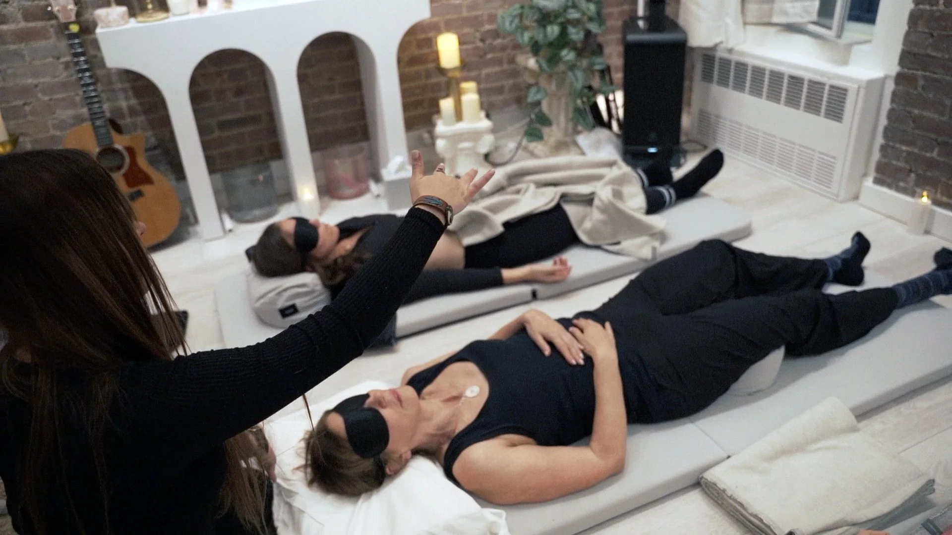 Two women lying on mats in a room with brick walls and candles, one with headphones, one resting with a hand on her stomach, and a woman standing nearby with her hand raised.