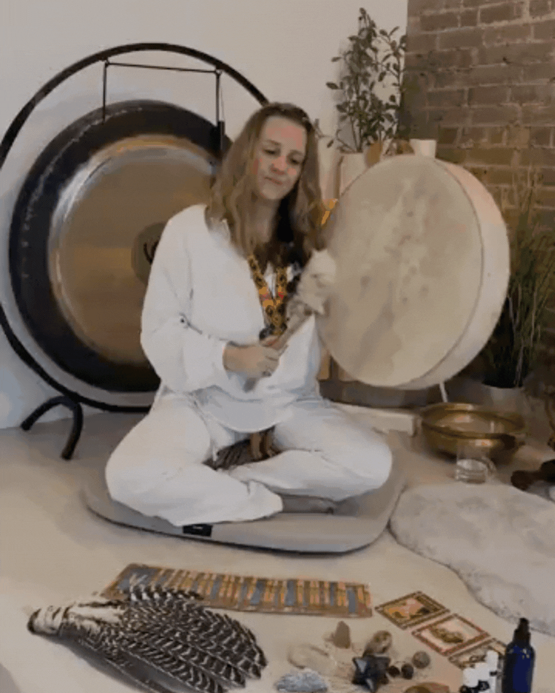 A woman in white pajamas sitting cross-legged on a cushion in front of a large drum, holding a drumstick, with other drums and talismans on the floor, and a washer in the background.