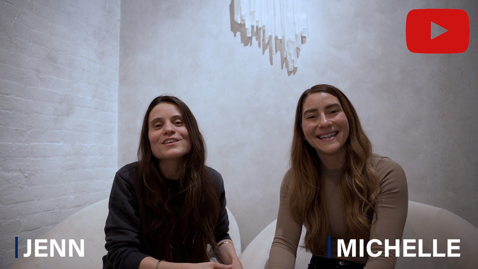 Two women, Jenn and Michelle, smiling and sitting on a white couch in a modern, minimalistic room with a white brick wall and an abstract light fixture overhead.