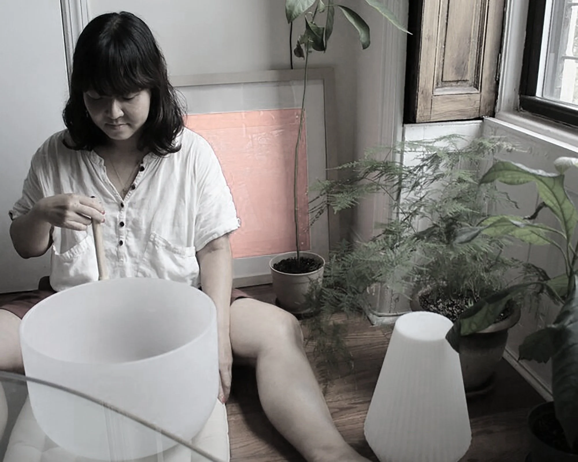 A woman sitting on the floor with her legs spread apart, playing a white handpan instrument with her eyes closed. She is wearing a white button-up shirt and is surrounded by plants and lamps near a window.