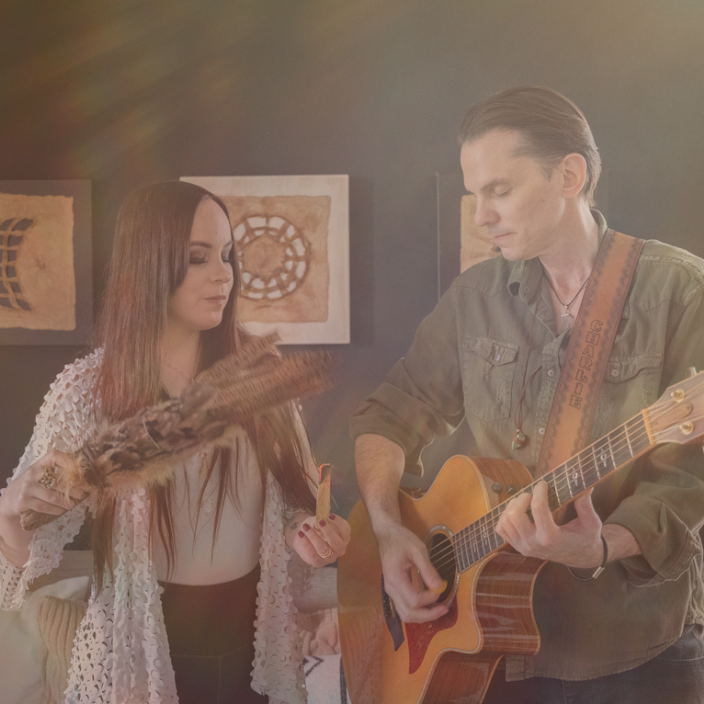 A woman holding a feathered fan and a man playing an acoustic guitar, standing in a room with framed artwork on the wall behind them.