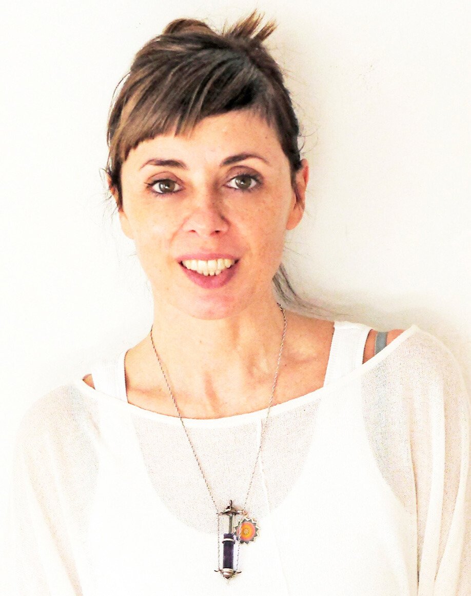 A woman with short, layered brown hair and light complexion smiling at the camera, wearing a white top with exposed shoulder strap and a necklace with a large, purple pendant.