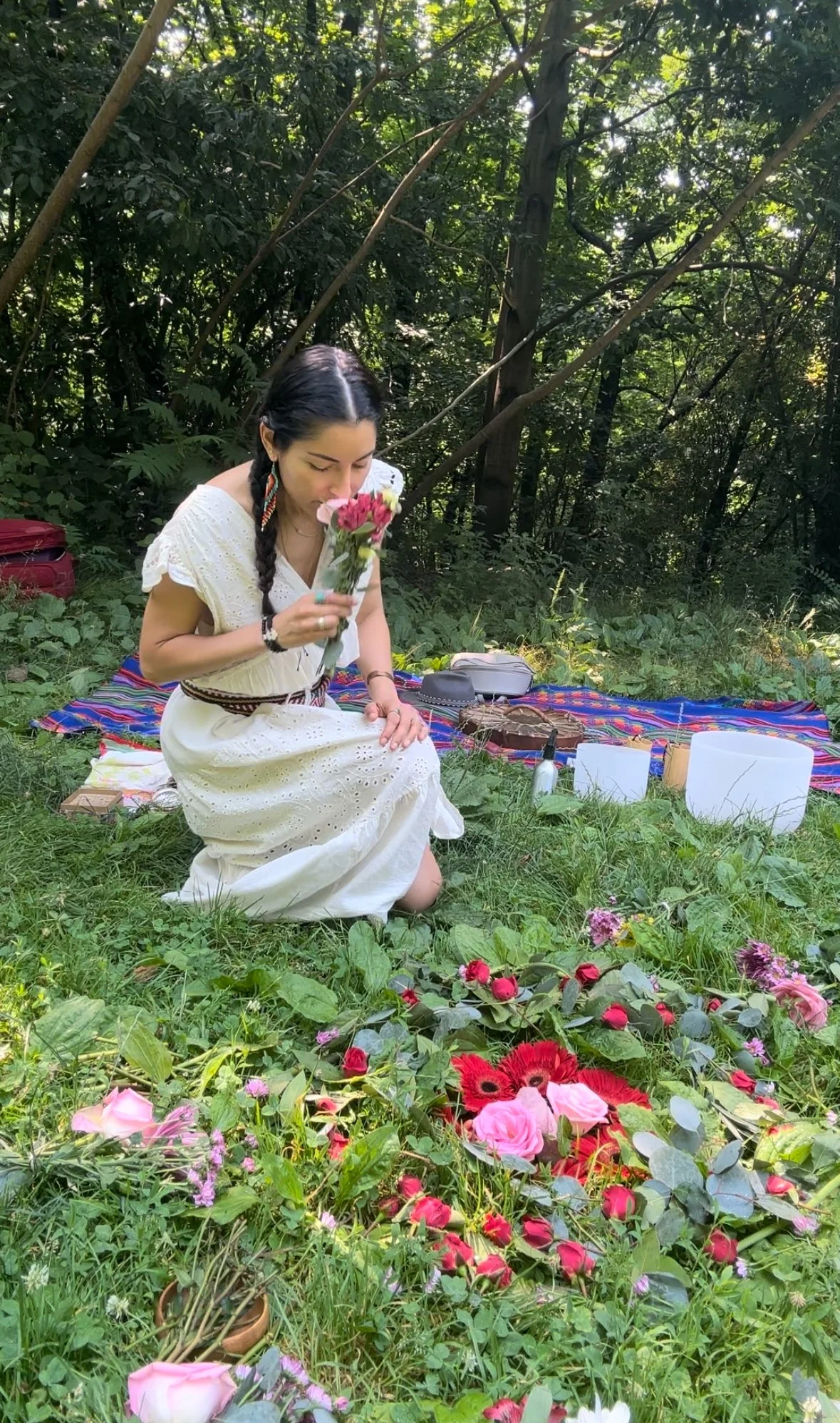 A woman dressed in a white dress with braided hair, kneeling on grass with a colorful blanket and flower arrangements around her, holding a flower bouquet and smelling it in a lush, green forest.
