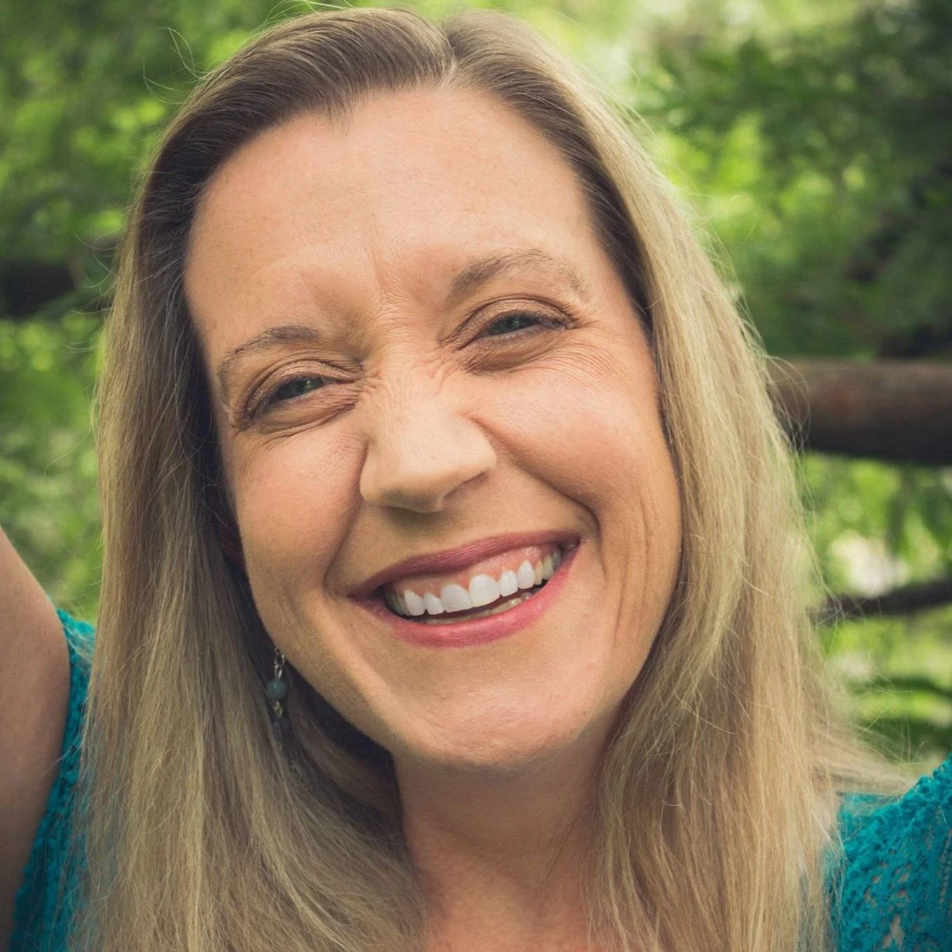 A woman with blonde hair smiling outdoors with greenery in the background.