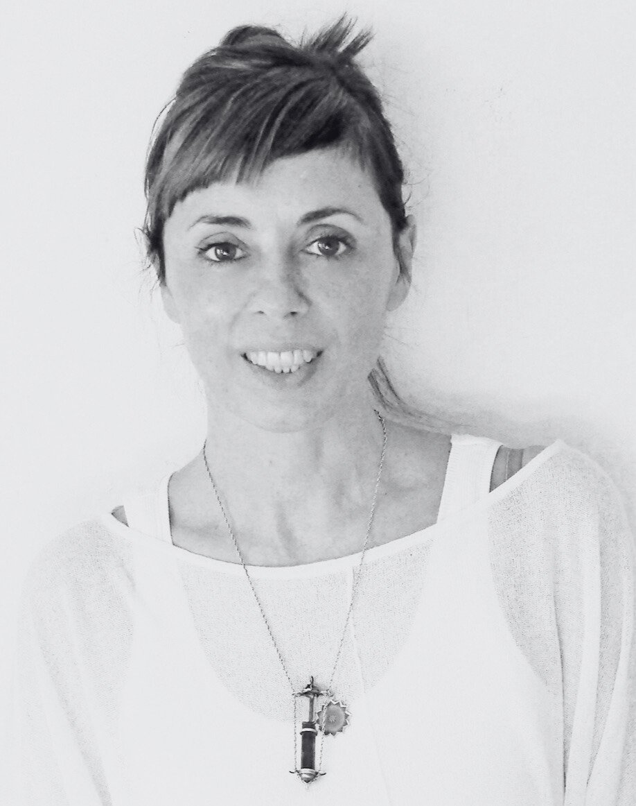 A woman with short, layered hair smiling, wearing a white top and a necklace with a key pendant, posing against a plain background.