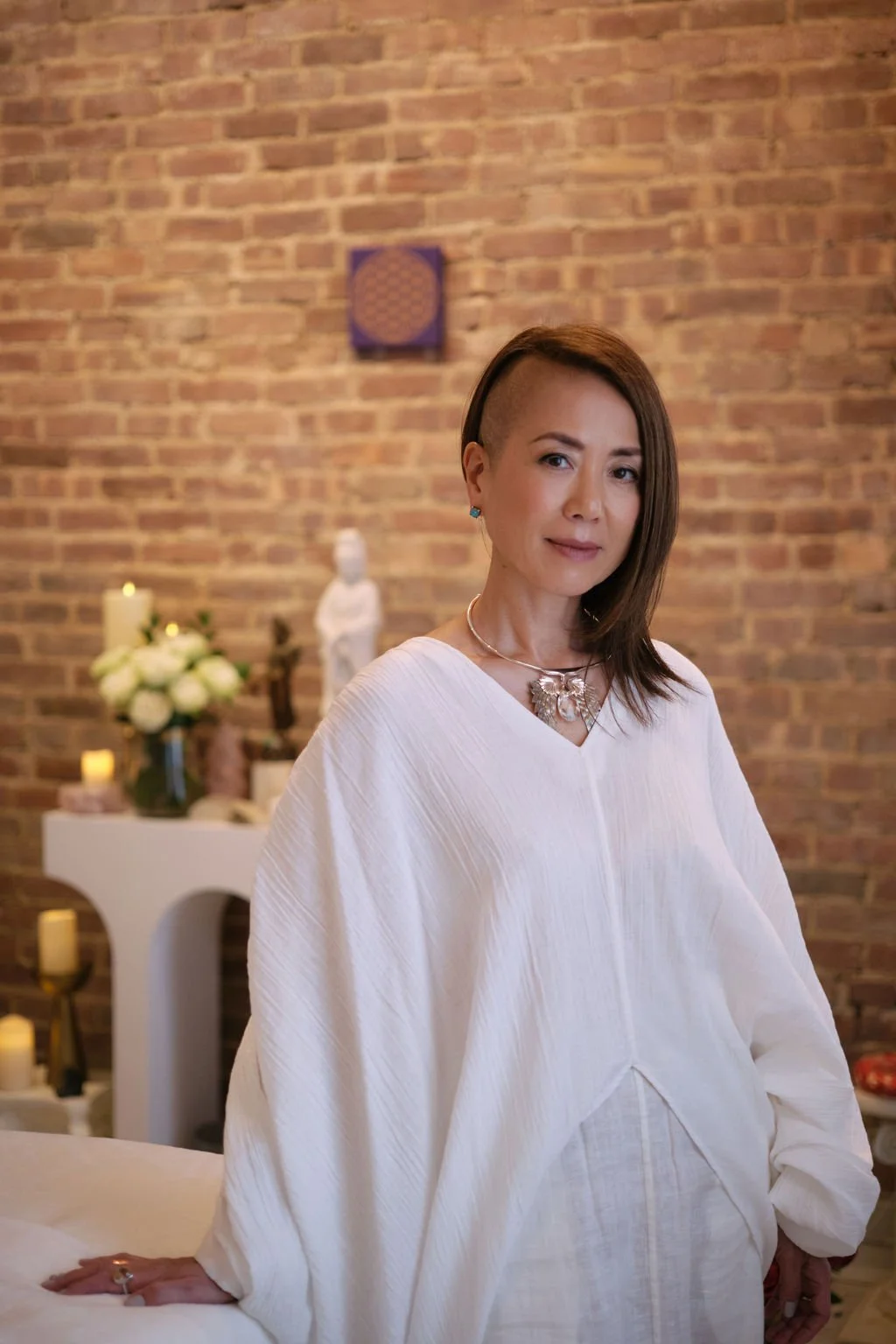 A woman with a partial undercut hairstyle and long brown hair, wearing a white outfit with jewelry, standing in a room with a brick wall and decorated altar or table in the background.