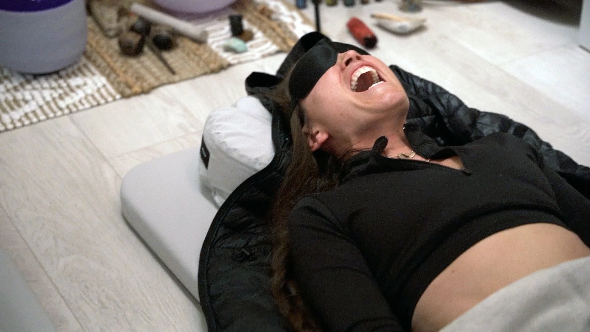 A woman lying on her back on a bed, laughing and wearing an eye mask and a black top.