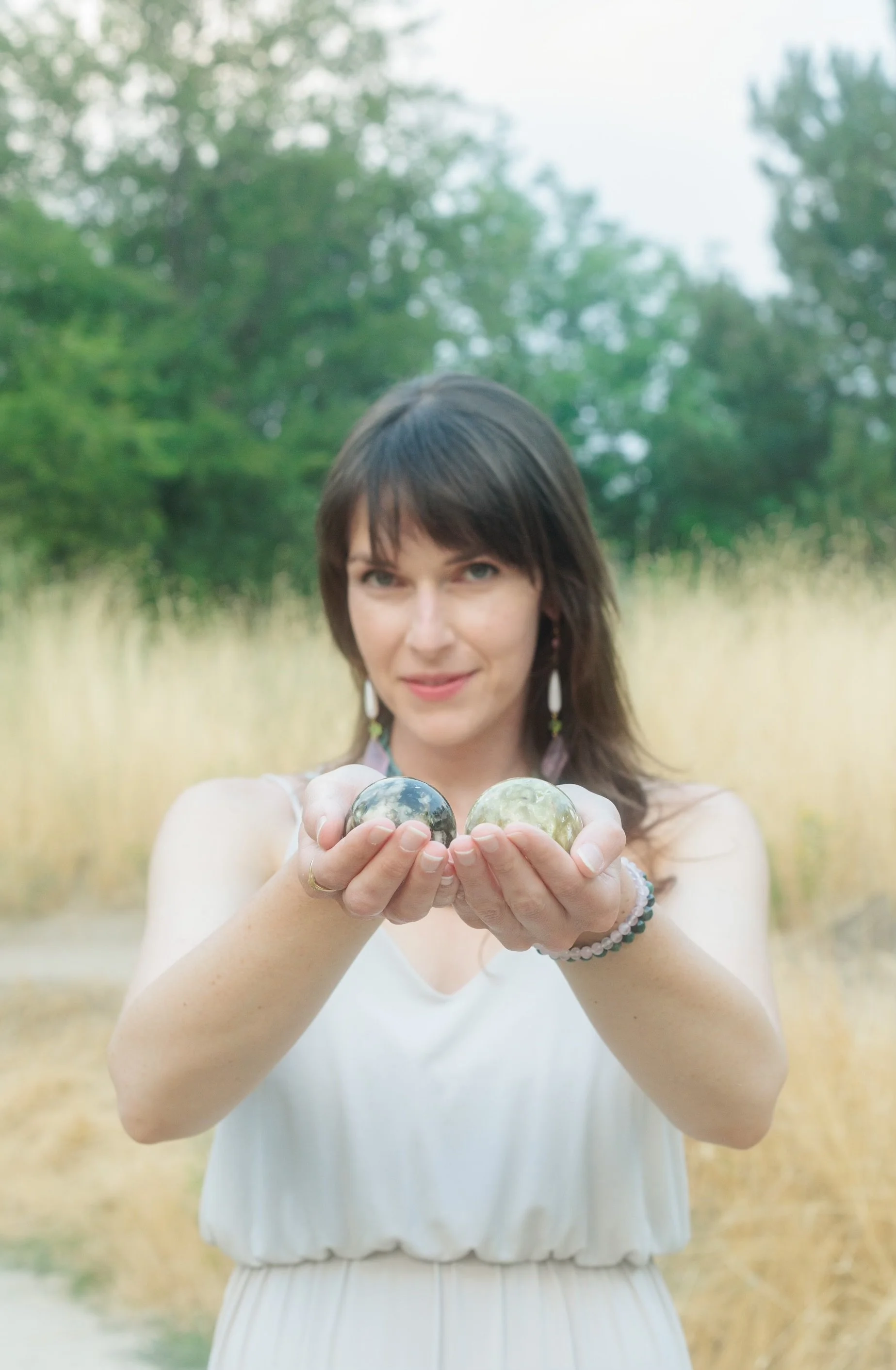 A woman with dark hair and earrings holding two shiny stones or marbles outdoors in a field with trees in the background.