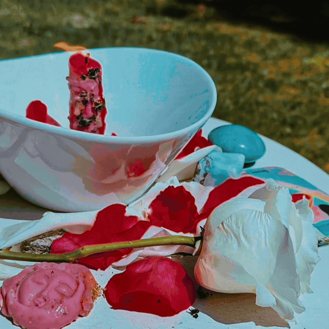 A white bowl with a pink pastry inside, surrounded by scattered rose petals, a white flower, and blue candies on a surface outdoors.