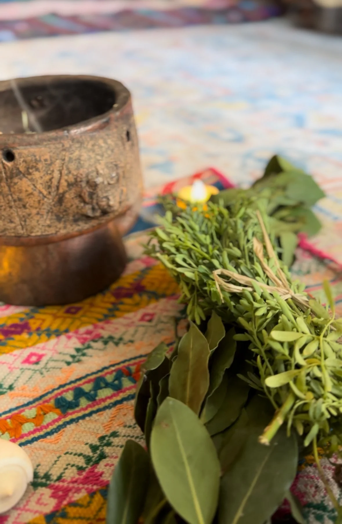A traditional altar with a burning incense holder, a bundle of fresh herbs, and a small candle on a colorful embroidered cloth.
