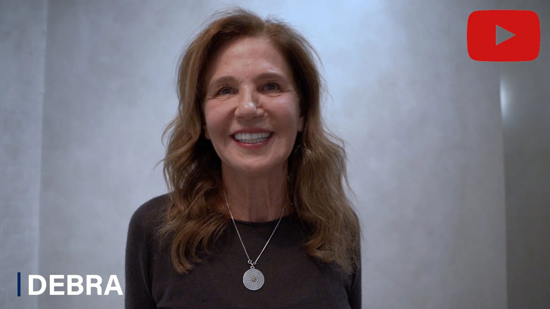 A smiling woman with brown, wavy hair wearing a black top and a silver necklace with a circular pendant, standing against a plain, light gray wall.