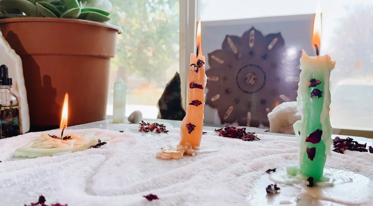 Woman in bohemian attire kneeling on floor surrounded by tarot cards, crystals, and candles, using a singing bowl, in a room with a brick wall, plants, and a large gong.
