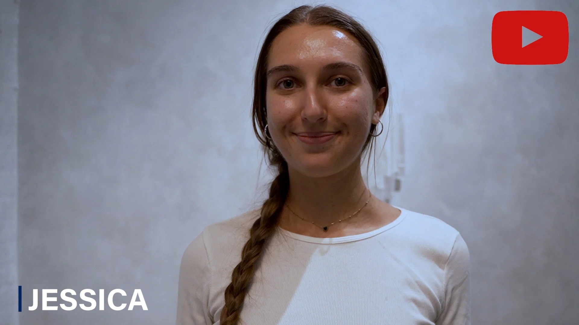 A young woman with brown hair in a braid, wearing a light-colored top and a delicate necklace, standing against a plain wall with a slight smile.
