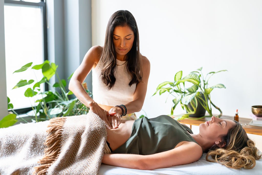 A woman receiving an abdominal massage from a therapist in a well-lit room with green plants.