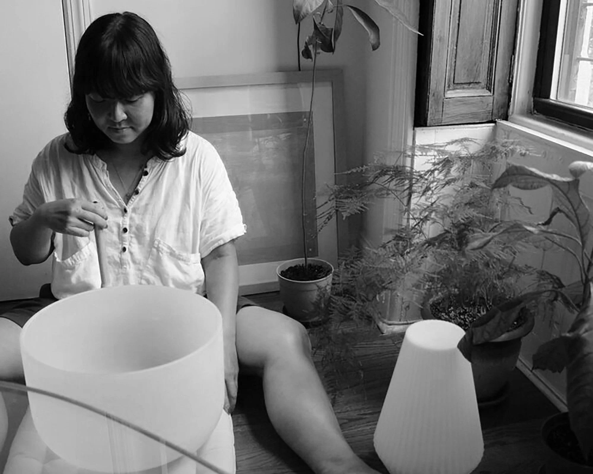A woman in a white shirt sitting on the floor in a room, with plants and lamp shades around her, near a window.