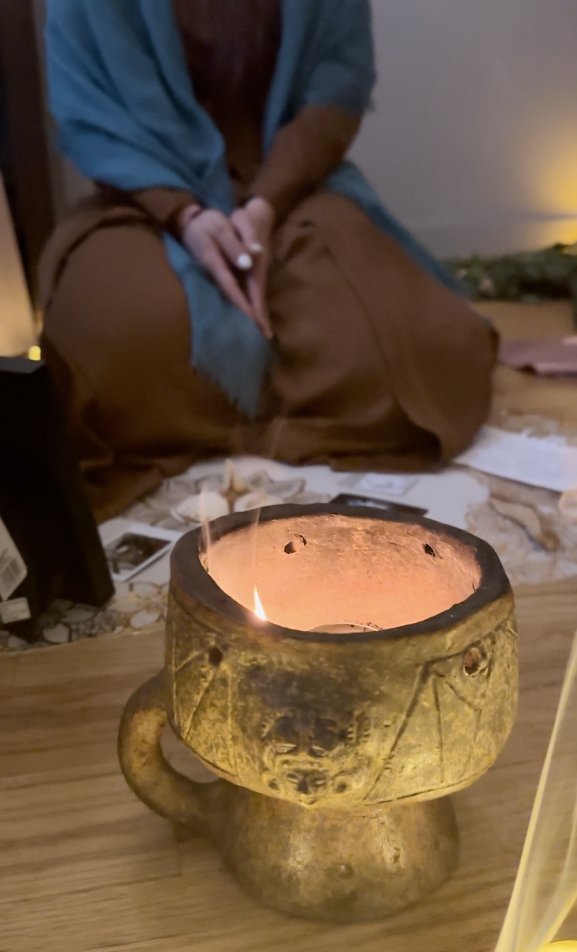 Close-up of a burning candle in an ornate, weathered holder on a wooden surface, with a person kneeling in the background. The person is dressed in a blue shirt and brown pants, with their hands clasped together.