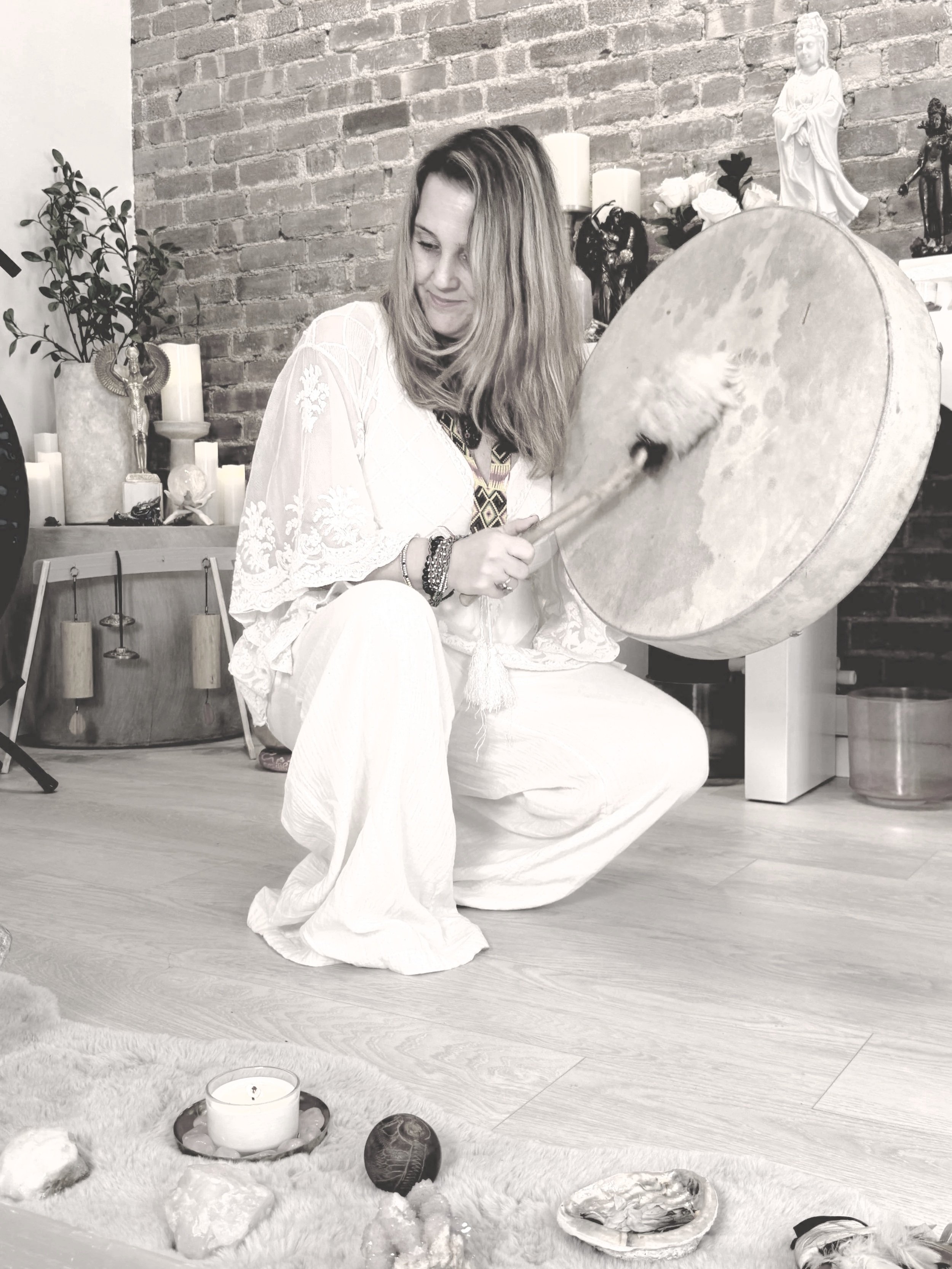 A woman kneeling on the floor and playing a large frame drum with a drumstick in a room decorated with candles, statues, and a brick wall.