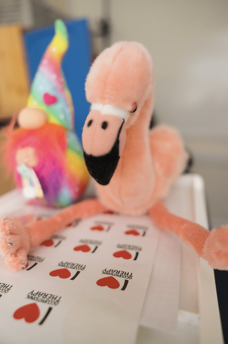 Stuffed flamingo toy with colorful gnome on a paper with "Occupational Therapy" and heart symbols.