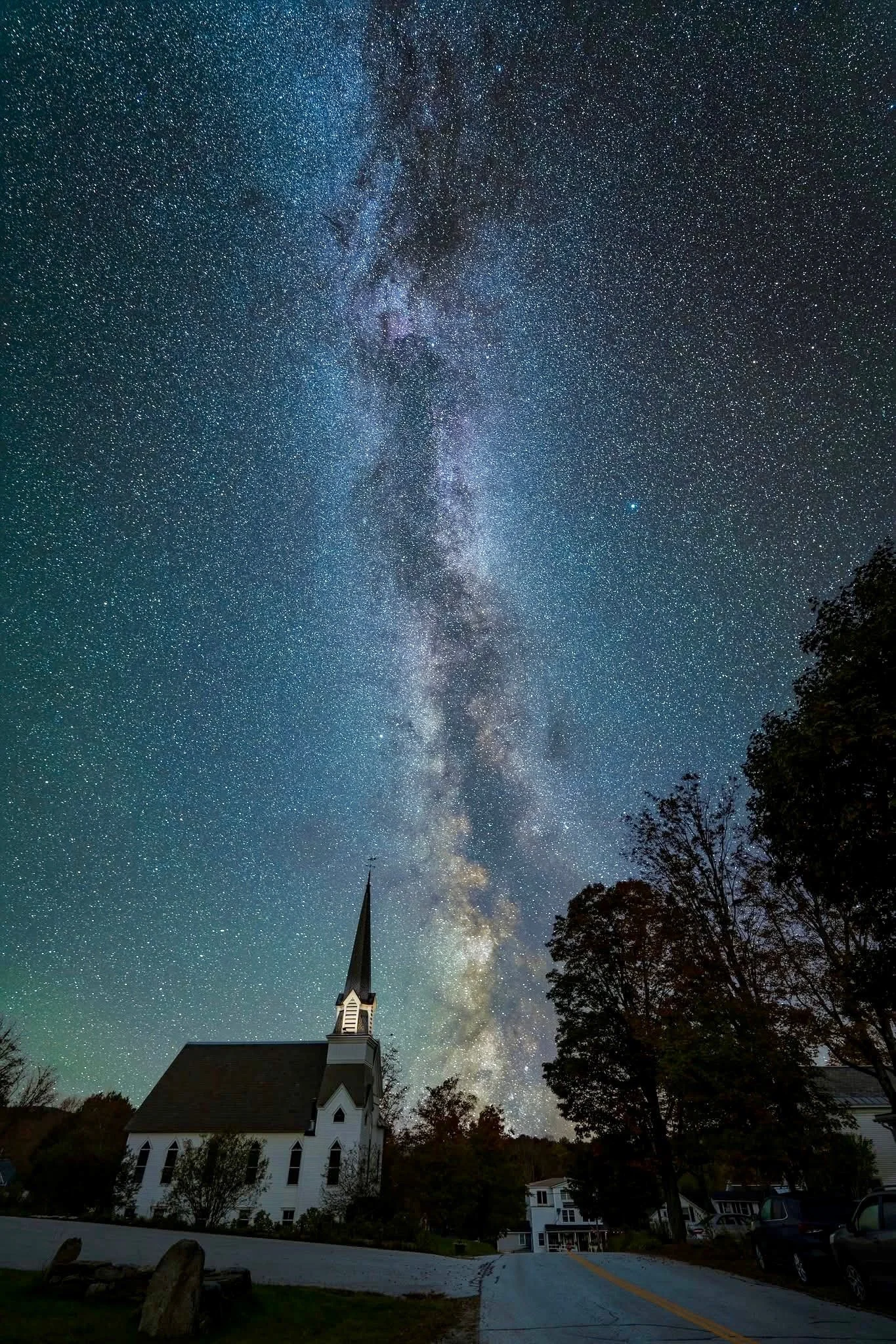 Night sky filled with stars and the Milky Way galaxy above a white church with a tall spire, trees, and parked cars.