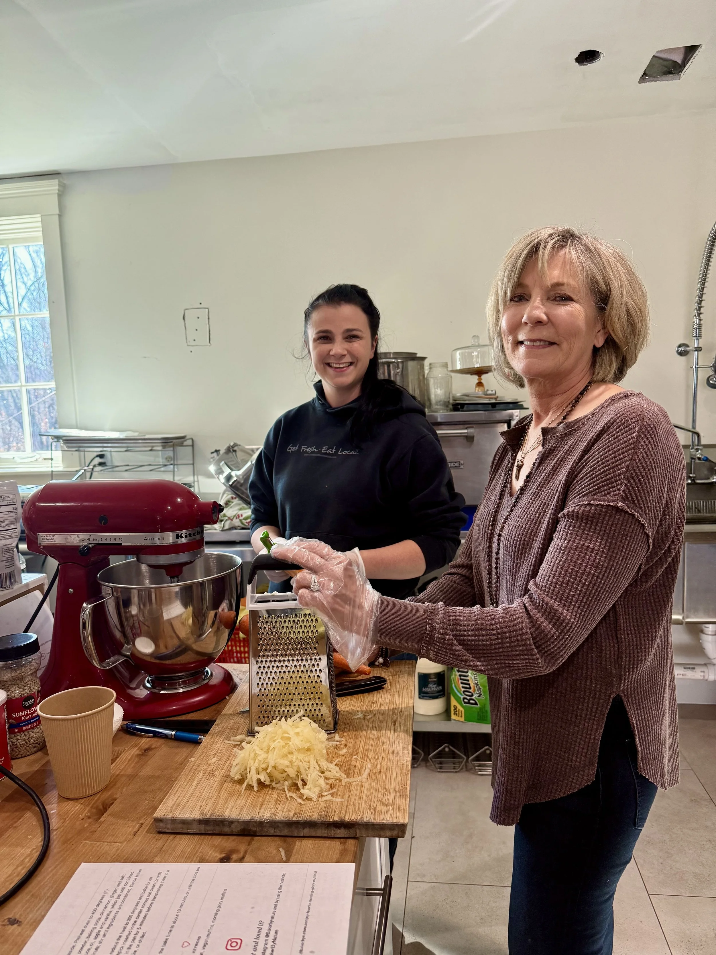 Two women in a kitchen preparing food; one is grating potatoes while the other stands behind a red stand mixer, smiling.