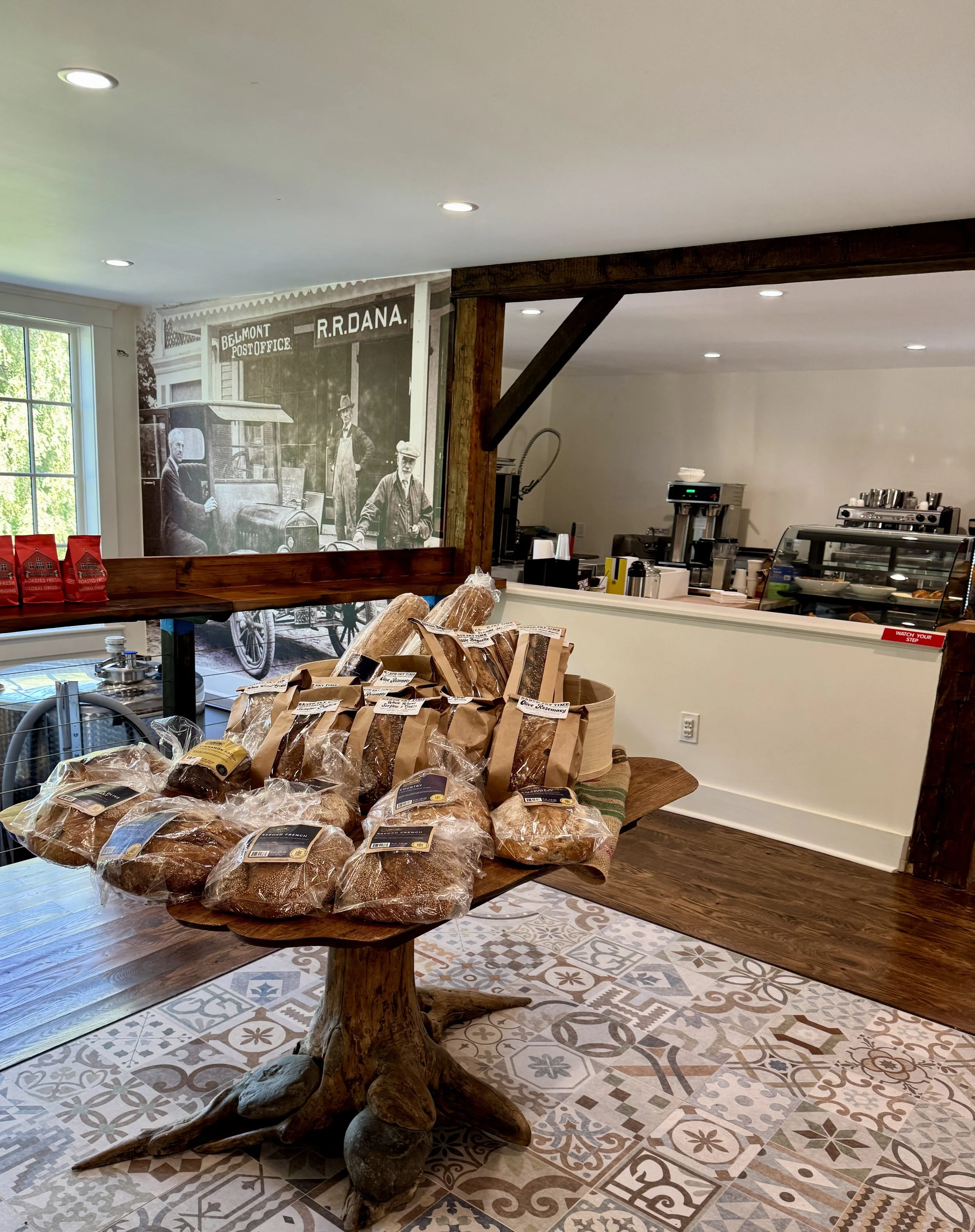 Display of bread on a rustic wooden table in a cafe or bakery, with a vintage black-and-white photo of the Belmont Post Office and R.R. Dana in the background