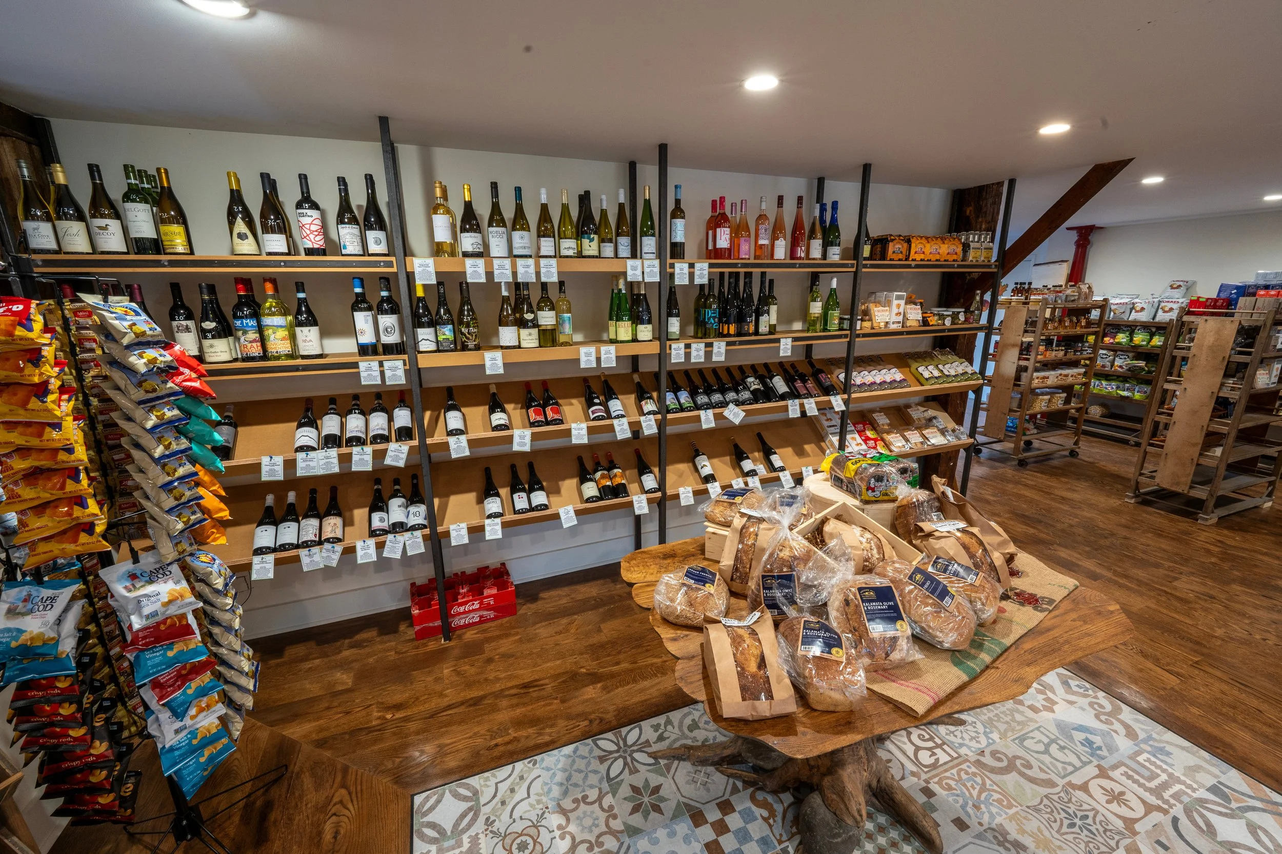Interior of a grocery store with wine bottles on shelves, snack bags on stand, and baked goods on a wooden table.