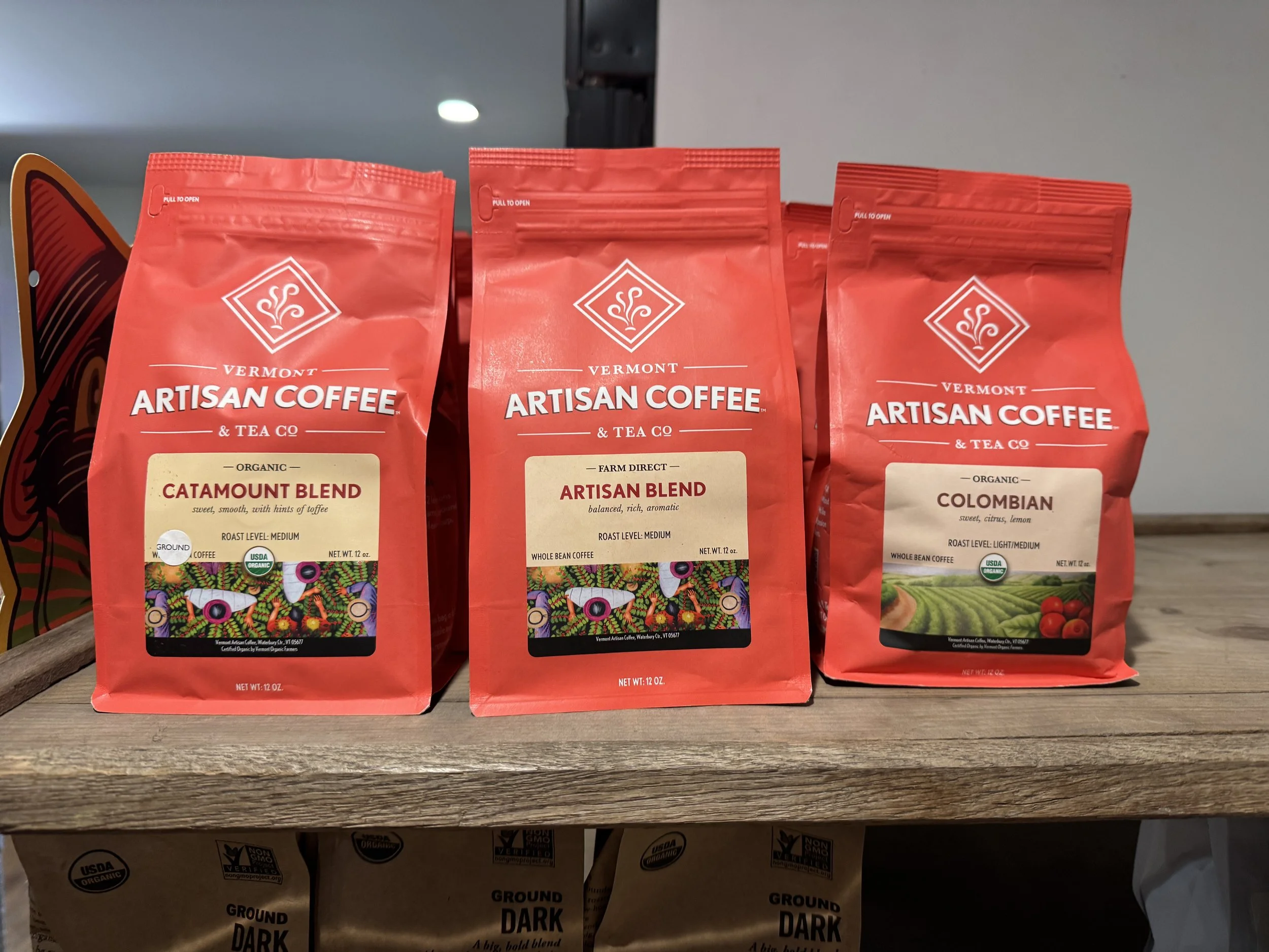 Three bags of Vermont Artisan Coffee on a wooden surface, with colorful designs and labels indicating blend types and organic certifications.