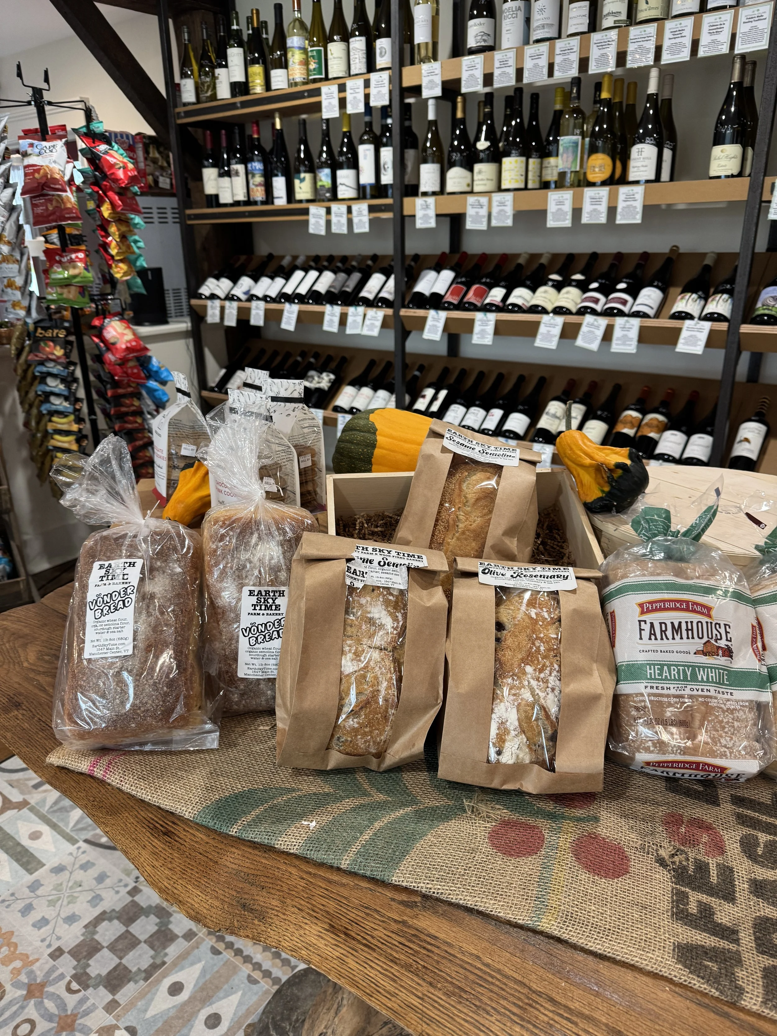 Gourmet bread and baked goods in paper and plastic bags on a wooden table, with a display of wine bottles on shelves in the background.