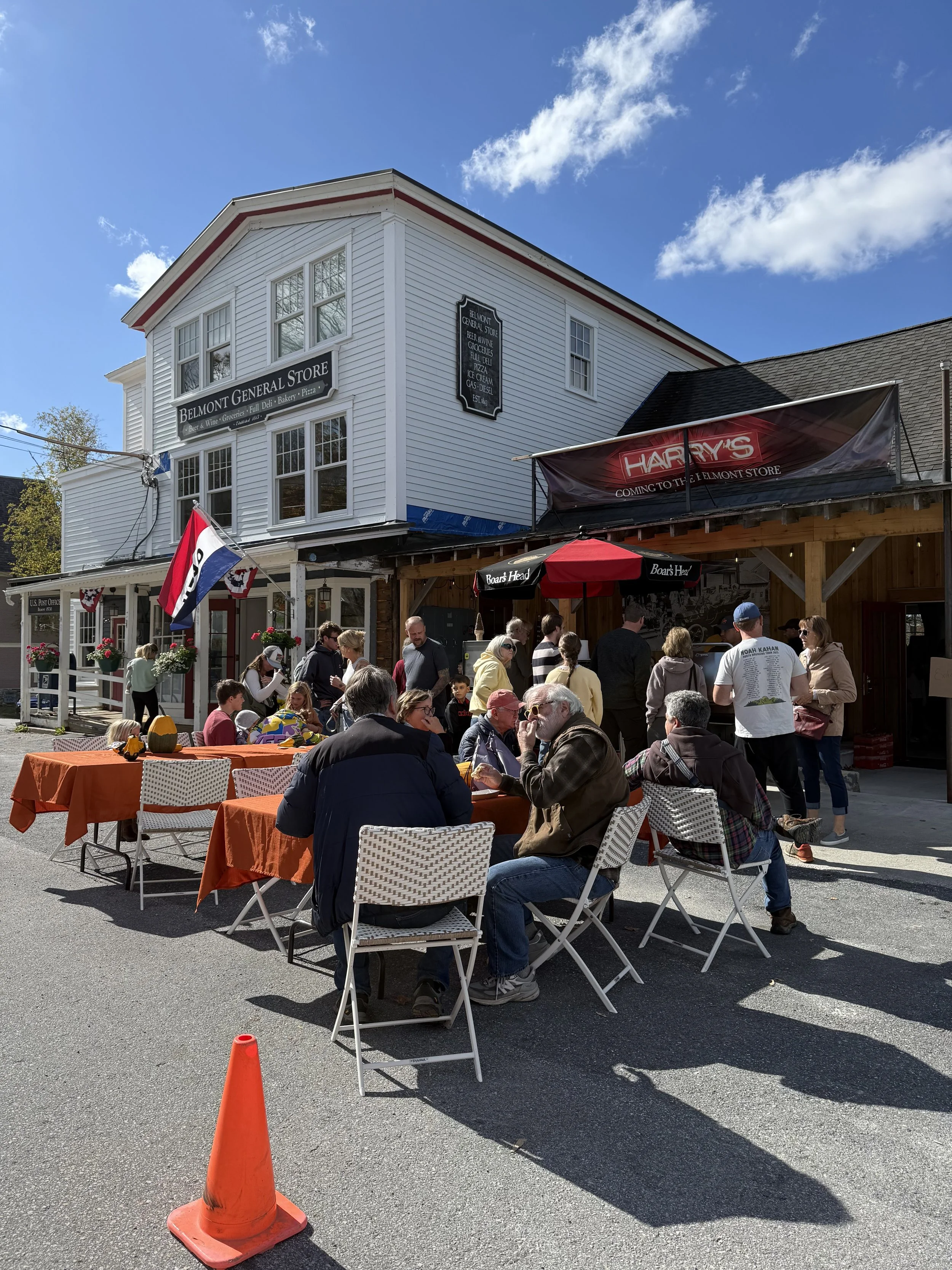 People sitting and standing outside a restaurant with orange tablecloths on tables, under a clear blue sky, in front of a white building with signs for a general store and Harry's.